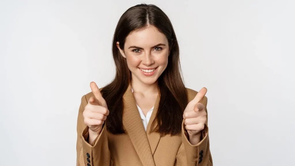 A woman in a brown blazer pointing both index fingers forward with a smile, against a white background.