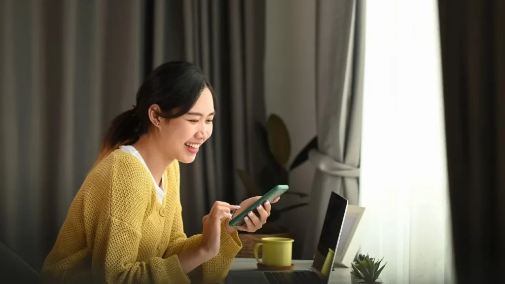 A person using a smartphone at a desk with a laptop.