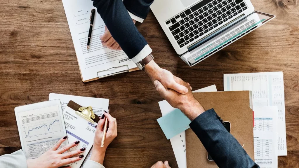 Two people shaking hands over a wooden table with documents, a laptop, and business papers.