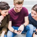 Three friends sitting on stone steps, smiling and using their smartphones together in a casual outdoor setting.