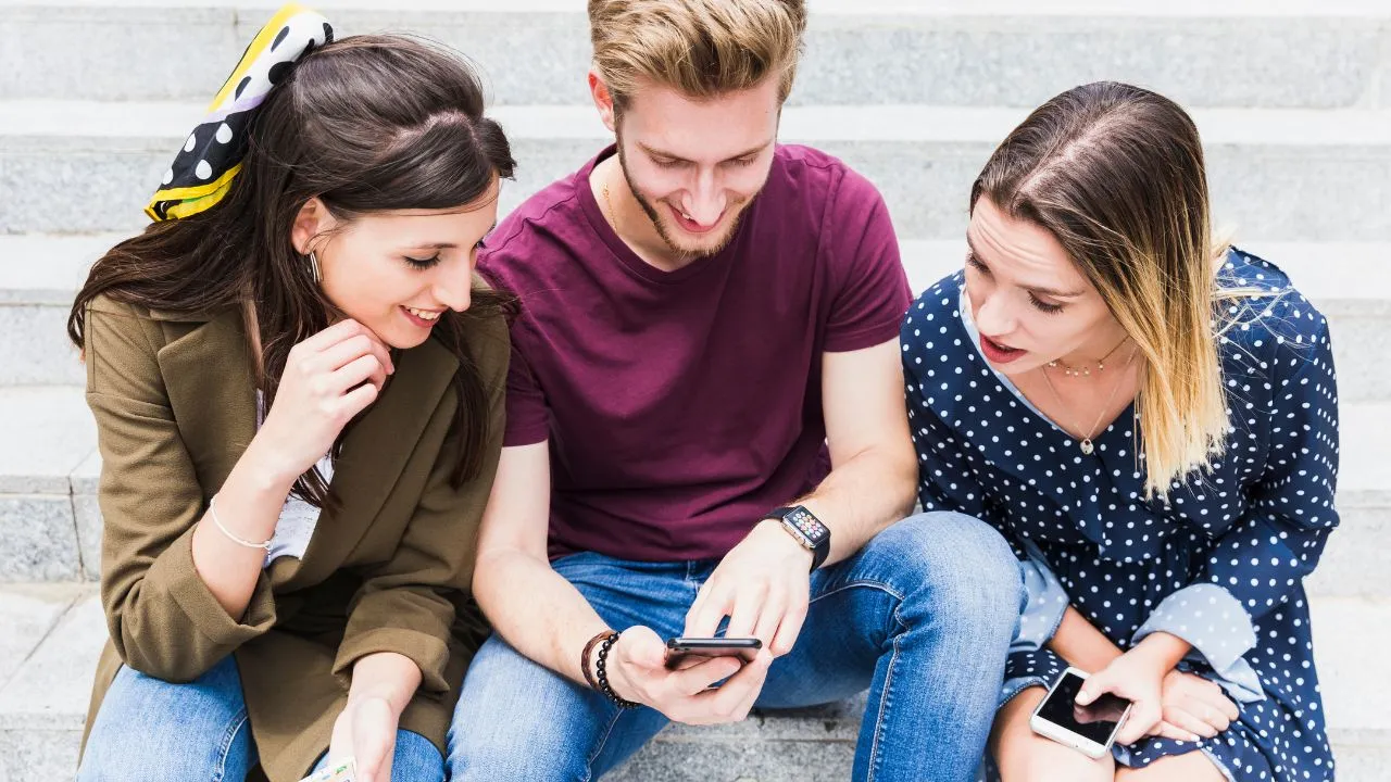 Three friends sitting on stone steps, smiling and using their smartphones together in a casual outdoor setting.