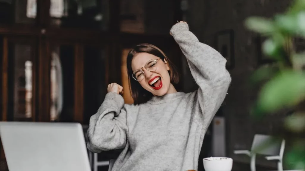 A woman in a gray sweater raising her arms in excitement while sitting at a desk with a laptop in a cozy indoor setting.