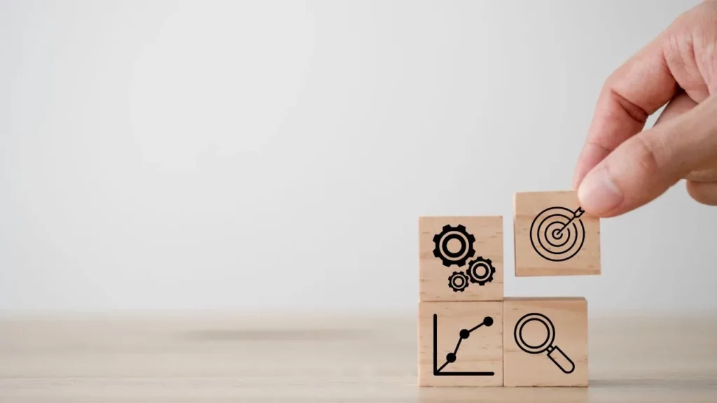 A hand arranging wooden blocks with icons representing goals, gears, growth charts, and a magnifying glass on a wooden surface.