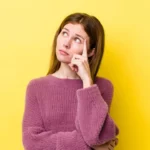 A woman with long brown hair wearing a pink sweater, standing against a yellow background, with her hand on her chin in a thoughtful pose.