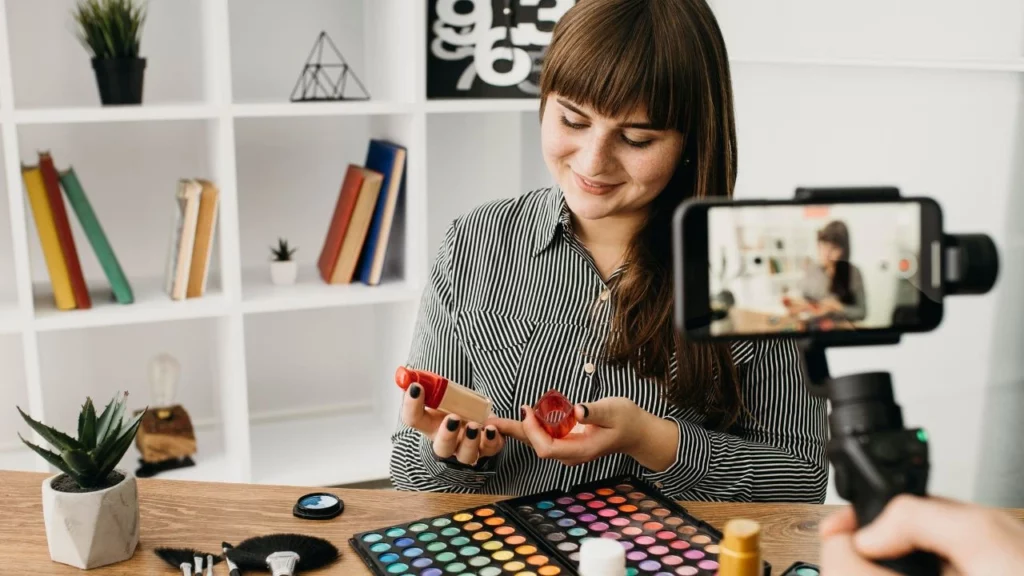 A woman with long brown hair recording a video, holding makeup products, with a colorful eyeshadow palette and brushes on the table in front of her.