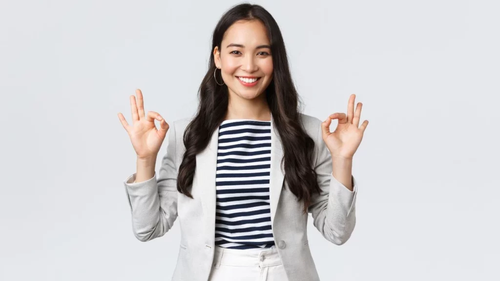A woman with long dark hair wearing a gray blazer and striped shirt, making an "OK" hand gesture with both hands against a plain background.
