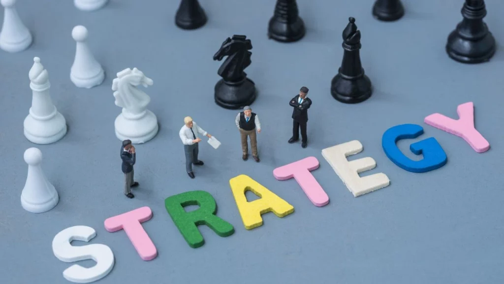 A chessboard with miniature figurines of people standing among chess pieces, and large colorful letters spelling "STRATEGY" in the foreground.