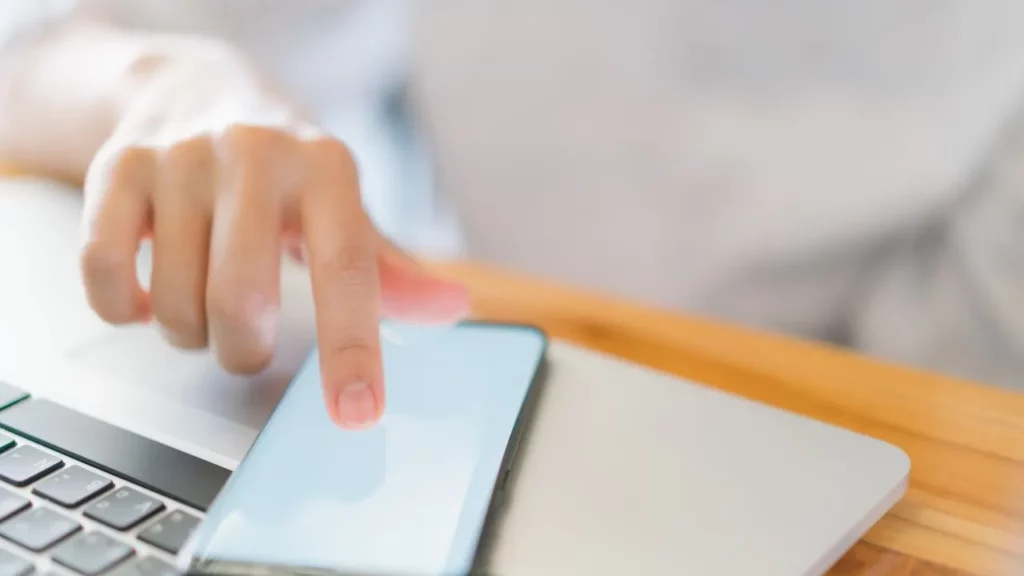 Hand tapping phone screen next to a laptop on a wooden table.