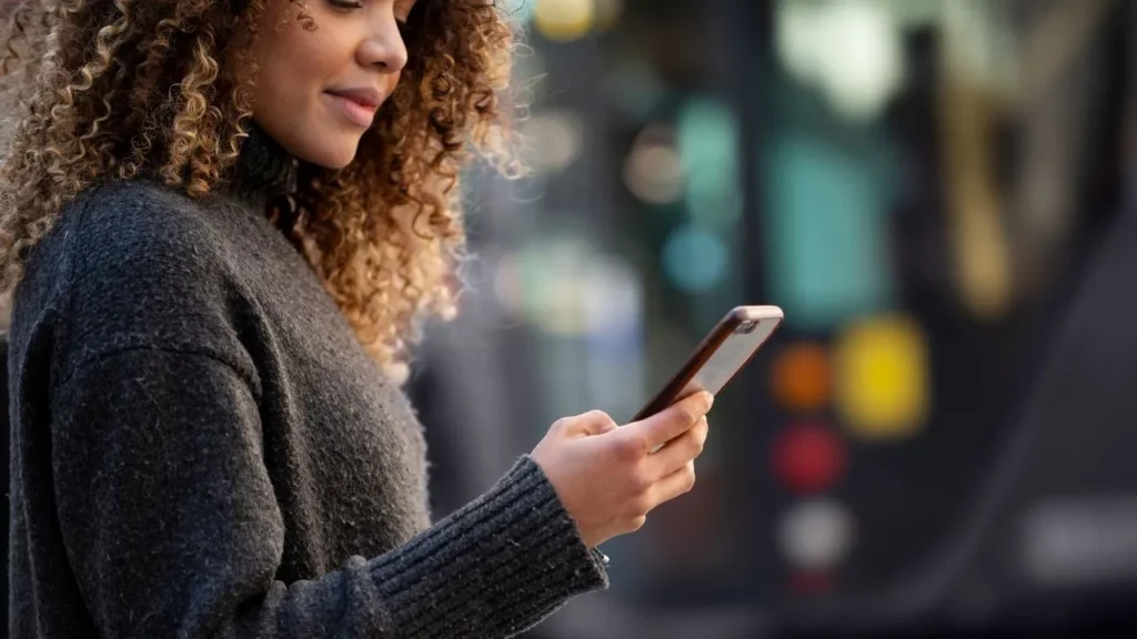 A woman with curly hair in a gray sweater holding a smartphone, looking at the screen, with a blurred urban background.