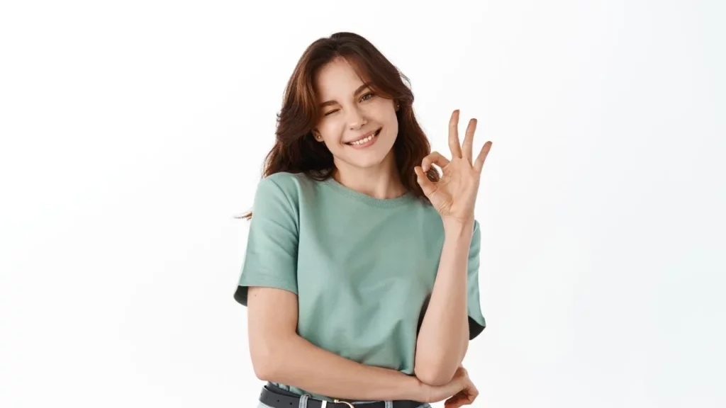 A woman in a green shirt giving an "OK" hand sign with her right hand, smiling and winking, against a white background.