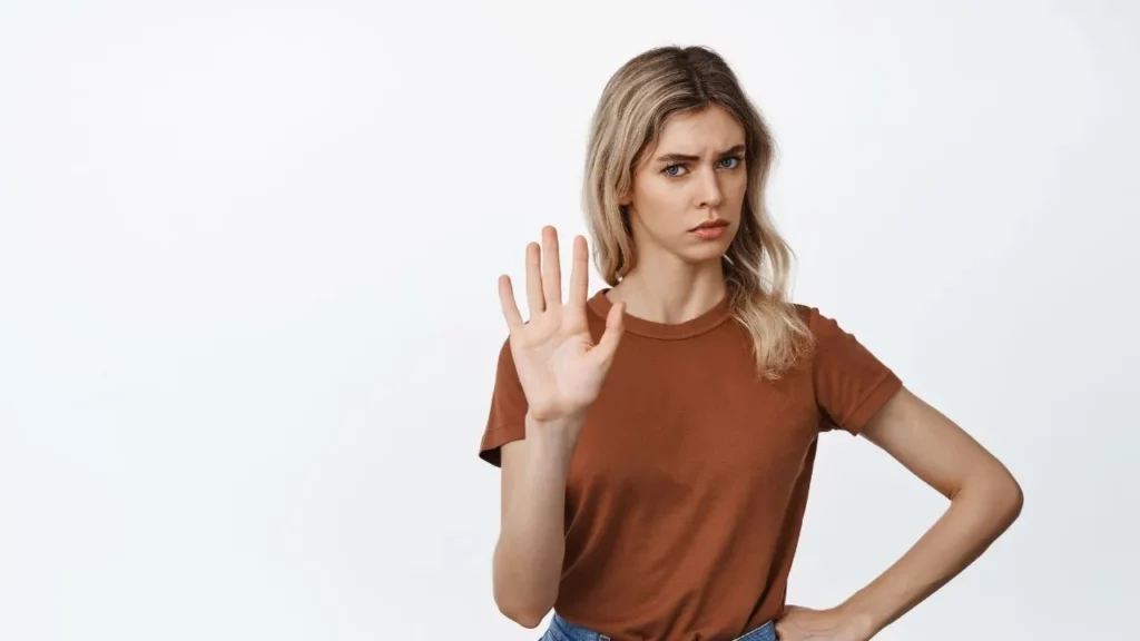 A woman in a brown shirt raising her right hand with an open palm, gesturing a stop sign, against a plain white background.