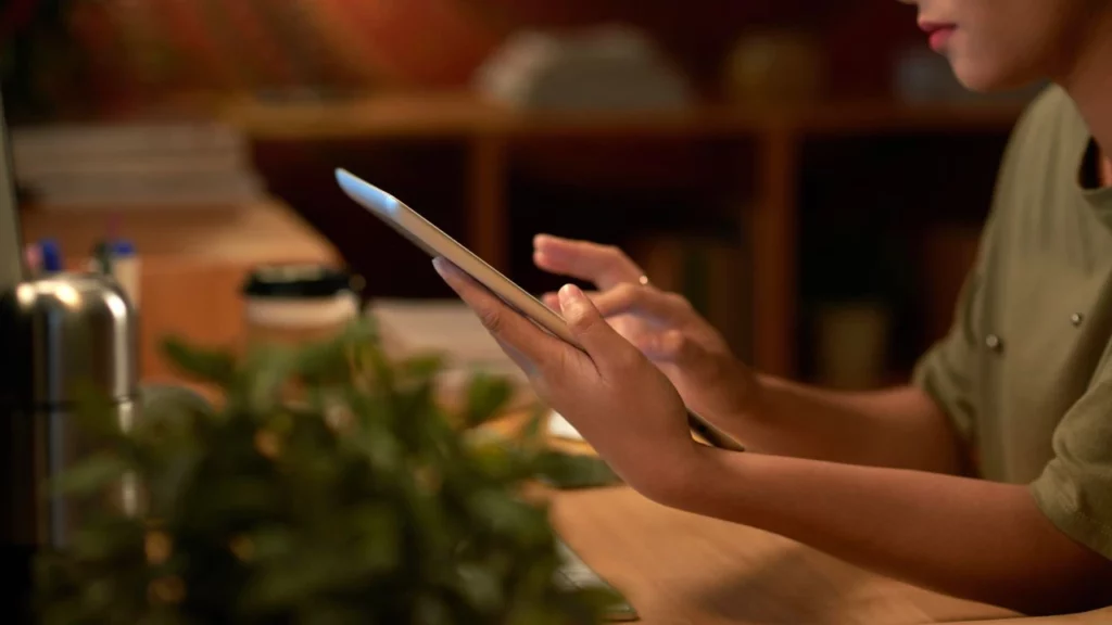 A side view of a person's hands holding and using a tablet in a dimly lit room, suggesting late-night work or browsing.