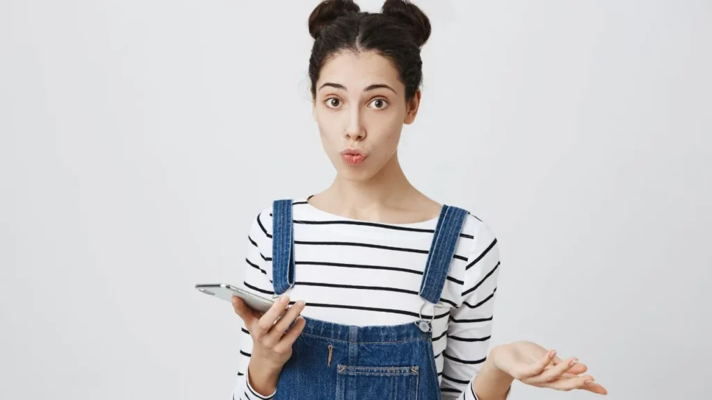 Confused young woman in overalls holding a tablet with a surprised expression.