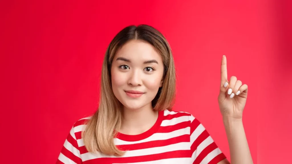 Smiling young woman in a striped shirt raising one finger against a red background.