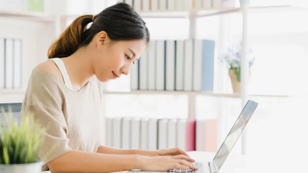 Woman working on a laptop in a bright office with bookshelves in the background.