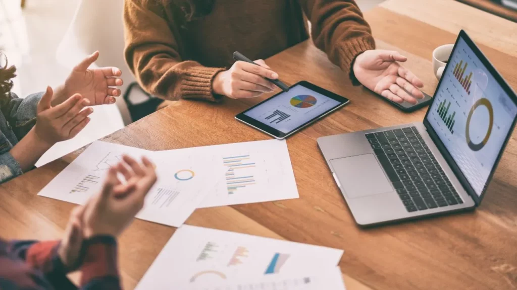 People analyzing graphs and charts on a tablet and laptop during a meeting.