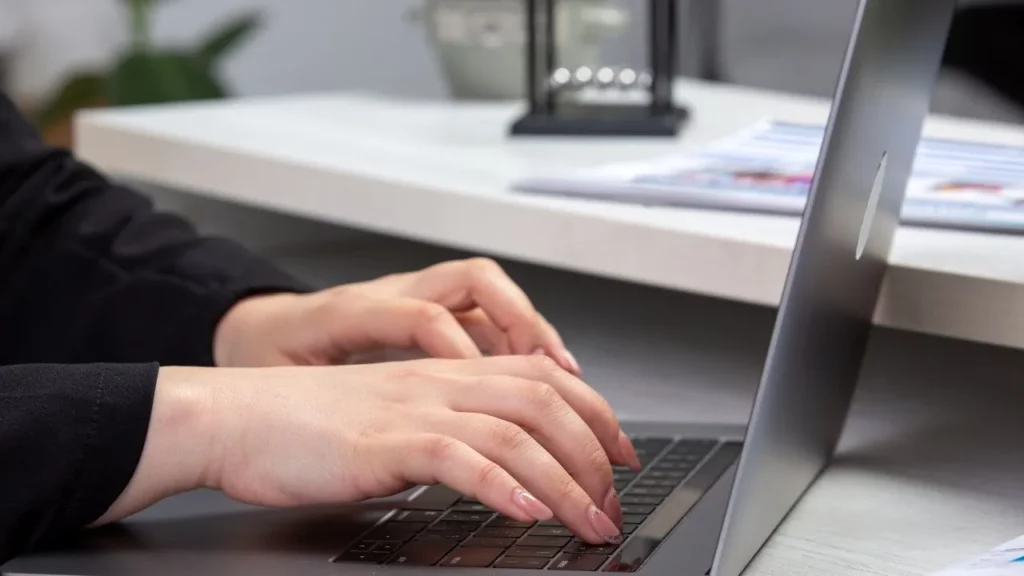 A person typing on a laptop at a desk with a white surface, a lantern, and some papers in the background.