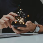 Person using smartphone with shopping icons above, laptop on desk.