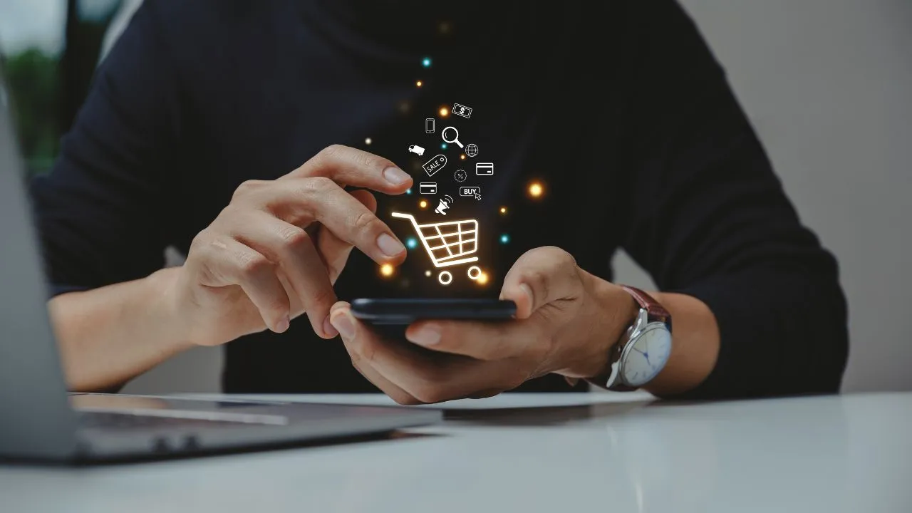 Person using smartphone with shopping icons above, laptop on desk.