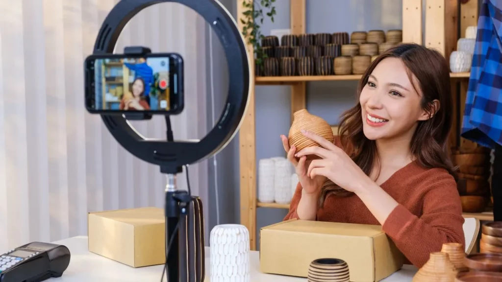 Woman live-streaming product demo with ring light, holding wooden vase in craft shop.