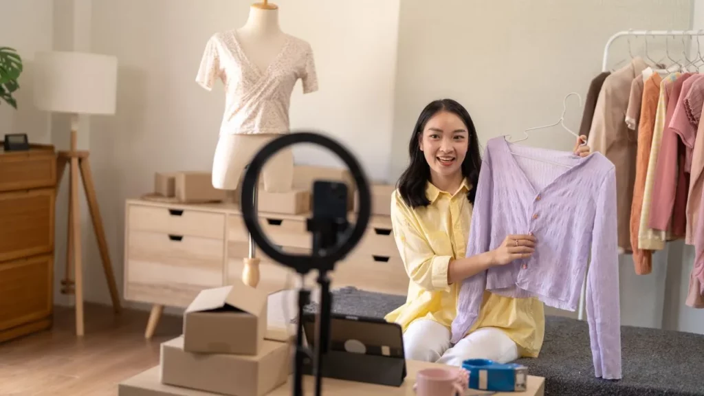 Woman live-streaming clothing haul, holding lavender blouse with ring light.