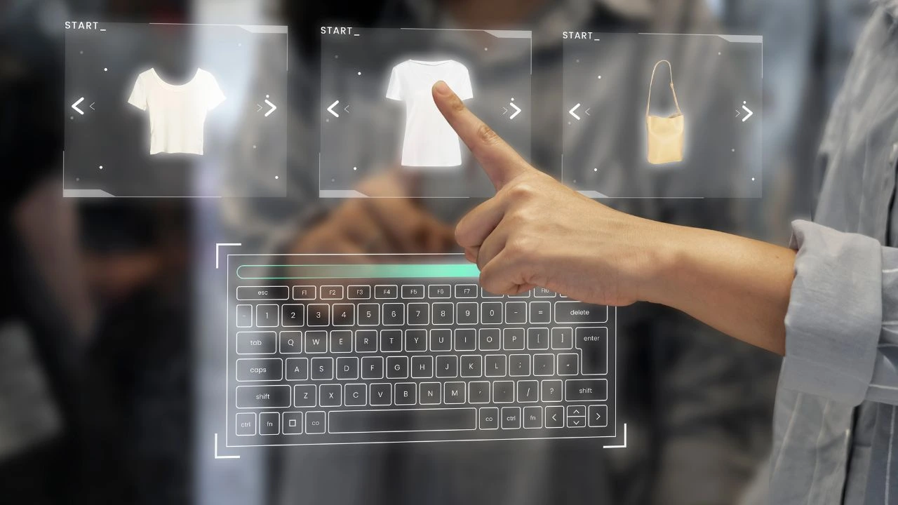 Hand interacting with a holographic interface showing clothing items like a white t-shirt and a beige handbag above a projected keyboard.