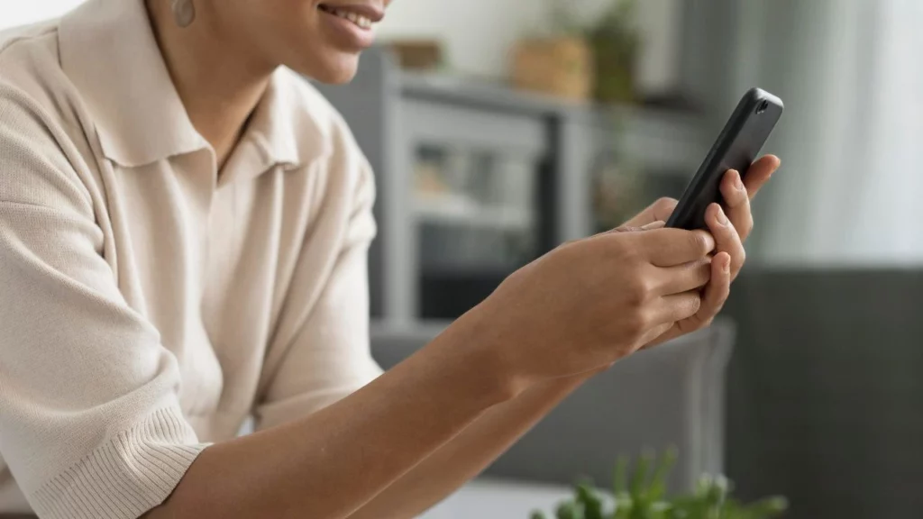 Person in a beige shirt smiling while using a smartphone in a modern living room setting.