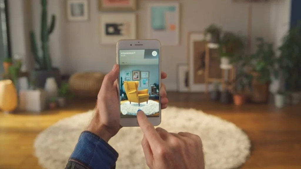 Person holding a smartphone displaying an augmented reality view of a yellow armchair placed in a living room with a rug and wall art.