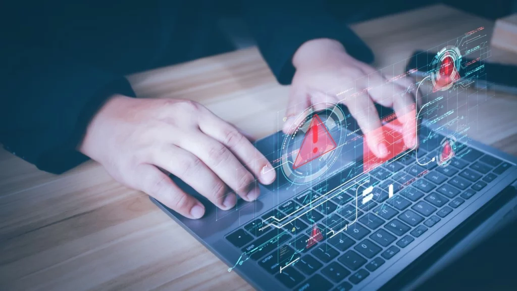 A person typing on a laptop with holographic warning alerts and red exclamation icons floating above the keyboard, indicating a cybersecurity threat.