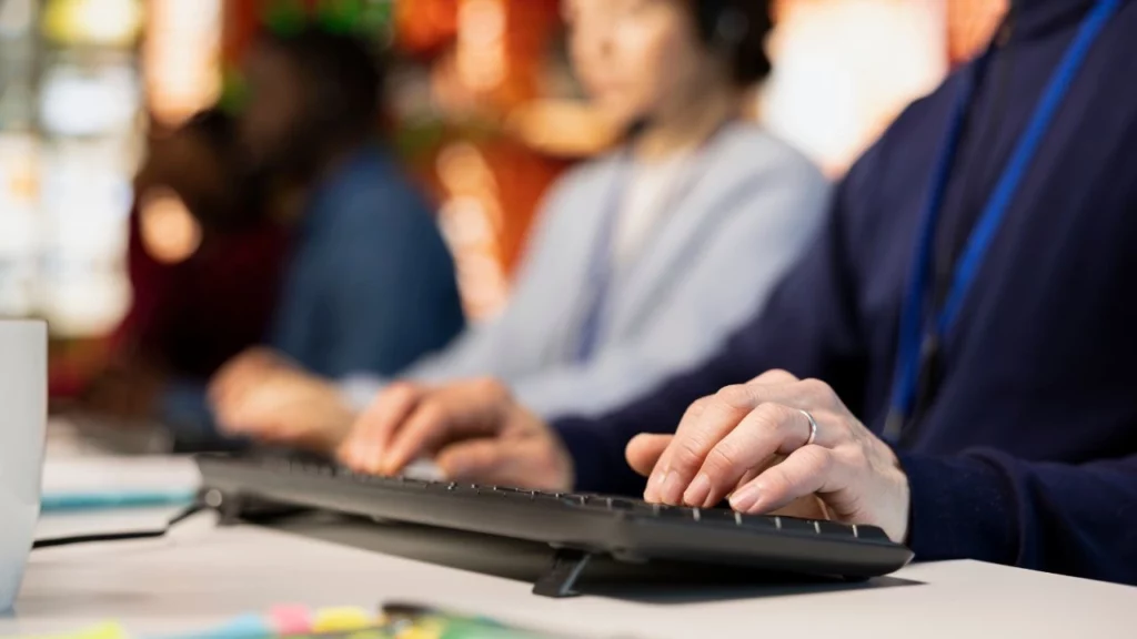 A person types on a keyboard in an office setting, with blurred colleagues working in the background.