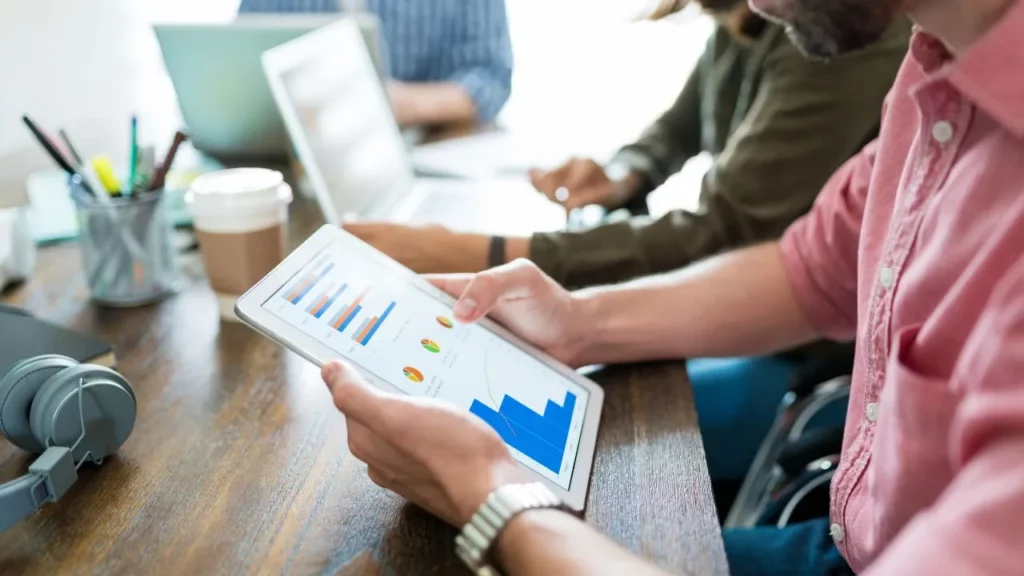 A person in a pink shirt holds a tablet displaying colorful bar graphs, pie charts, and line graphs, while others work on laptops in the background.