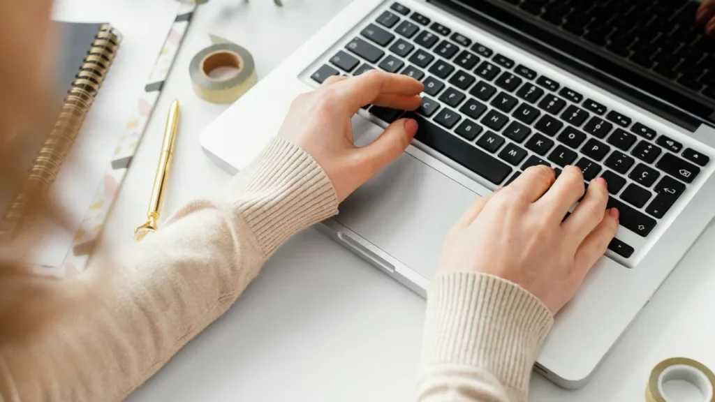 Close-up of hands typing on a silver laptop surrounded by notebooks and washi tape, depicting a productive writing workspace.