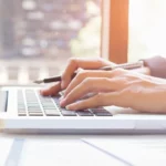 Close-up of a person typing on a silver laptop in a sunlit room.