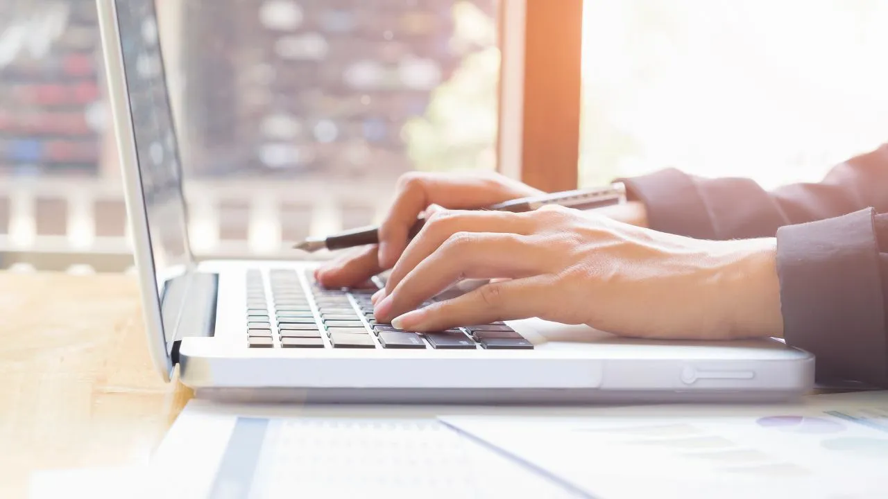 Close-up of a person typing on a silver laptop in a sunlit room.