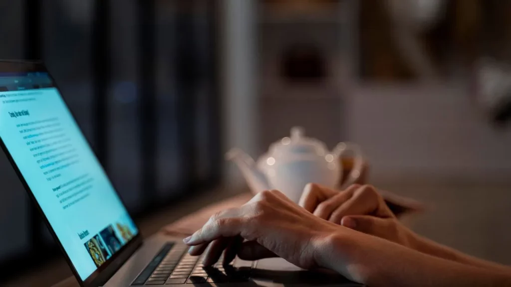 Hands typing on laptop in dim room with teapot in background.