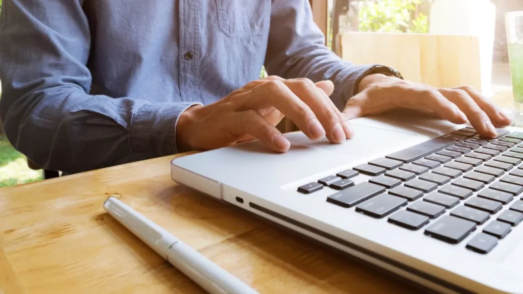 Close-up of hands typing on a silver laptop at a wooden desk in a sunny room.