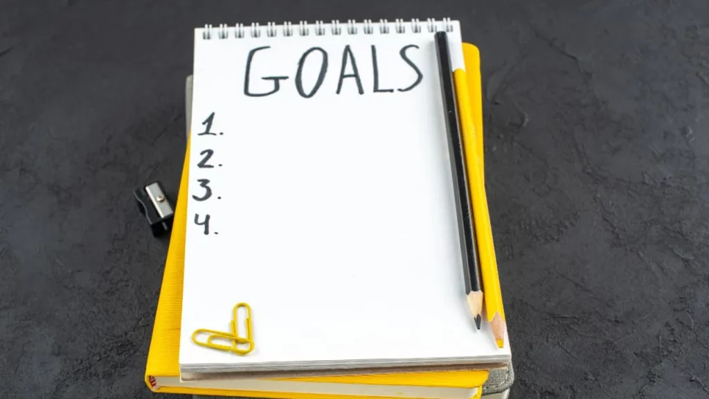 Top view of a blank notepad labeled "GOALS" with numbered lines, pencils, a paperclip, and sharpener on a dark surface.