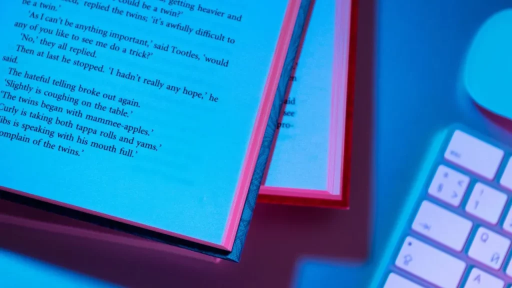 An open physical book with visible text on the pages placed next to a glowing laptop keyboard, illuminated in blue and purple tones, suggesting the contrast or combination of traditional reading and digital technology.