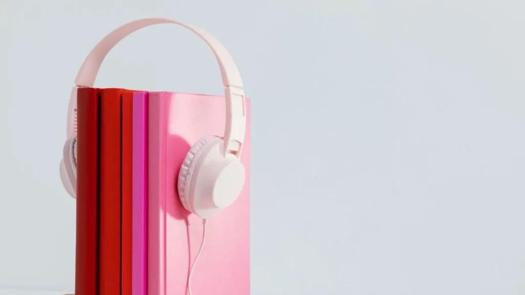 A pair of white over-ear headphones placed around a stack of colorful books with spines in shades of red, orange, and pink, against a plain light background. The image symbolizes audiobooks or listening to literature.