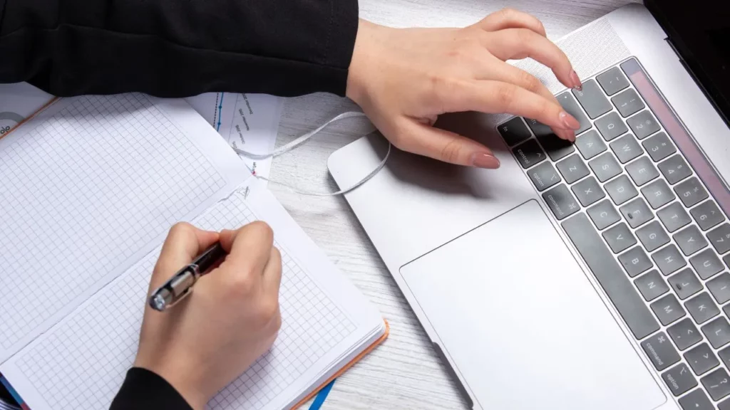 A person multitasking at a desk, typing on a silver laptop with one hand while writing notes in a grid notebook with the other hand.