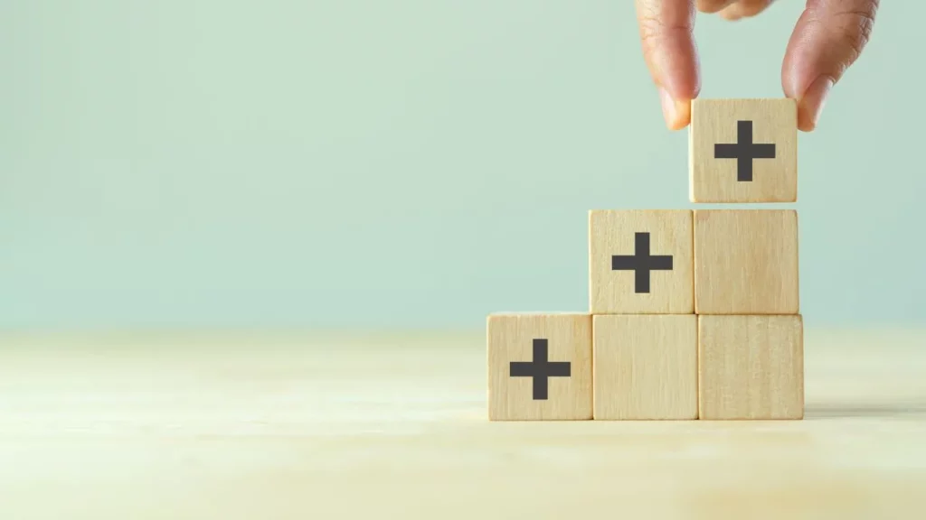 A hand placing a wooden block with a plus sign atop a small staircase of similar blocks.