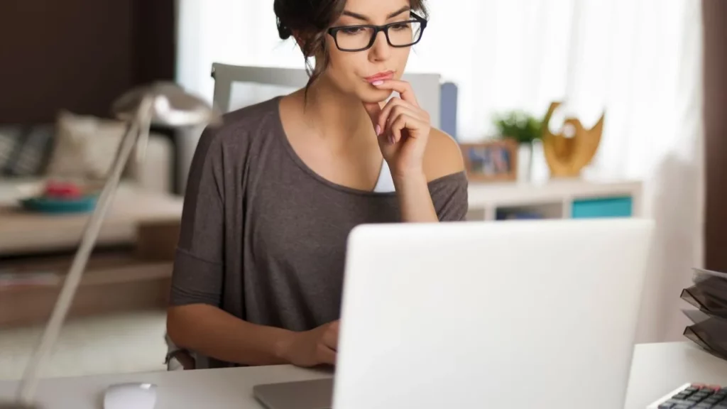 A woman with glasses looking at a laptop screen with a thoughtful expression.