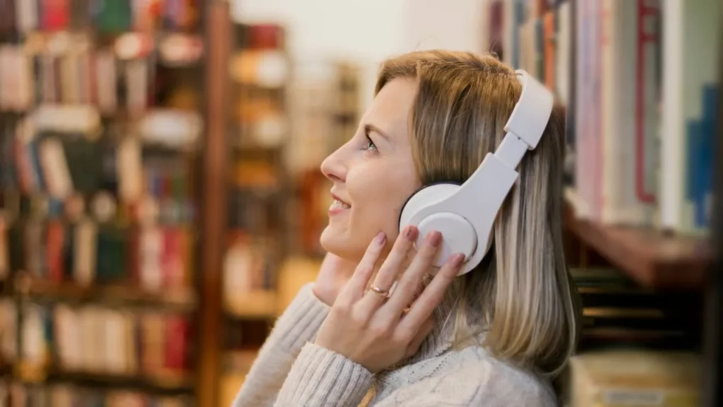 A woman wearing white headphones smiling in a library or bookstore.