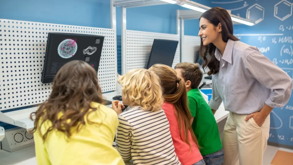 A smiling female science teacher in a modern classroom lab pointing at a large monitor displaying a brain scan while a group of excited young children gather closely around her, looking at the screen.