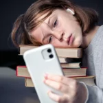 A young girl with freckles and a ponytail rests her head tiredly on a tall stack of colorful books while staring intently at her smartphone screen, wearing wireless earbuds.