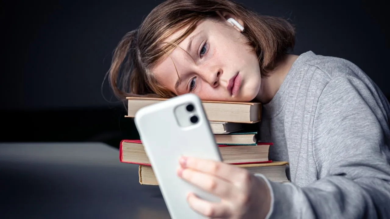 A young girl with freckles and a ponytail rests her head tiredly on a tall stack of colorful books while staring intently at her smartphone screen, wearing wireless earbuds.
