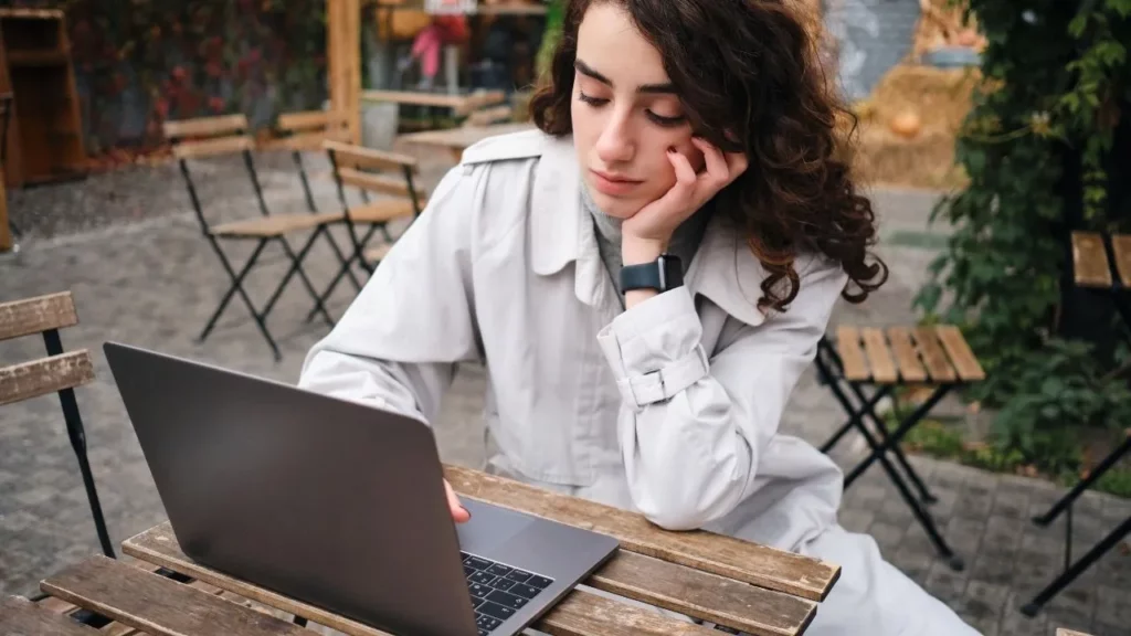 A young woman with curly hair and a thoughtful, slightly weary expression sits outdoors at a wooden table, resting her head on her hand while focused on an open laptop, wearing a light trench coat and smartwatch.