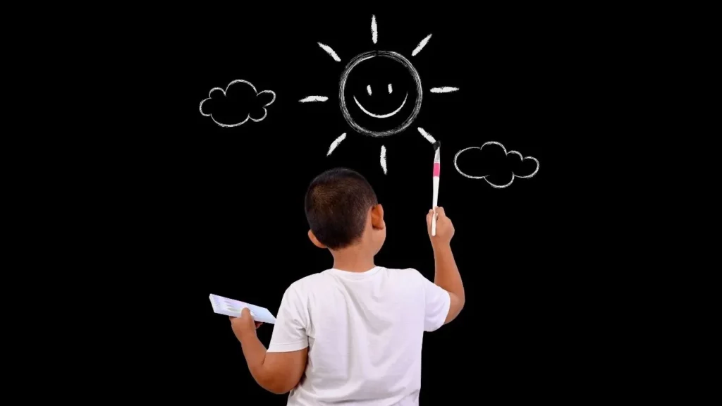 A joyful young boy stands in front of a blackboard, smiling as he paints a bright smiling sun with radiating rays, fluffy clouds, and a paintbrush in his hand against a dark background.