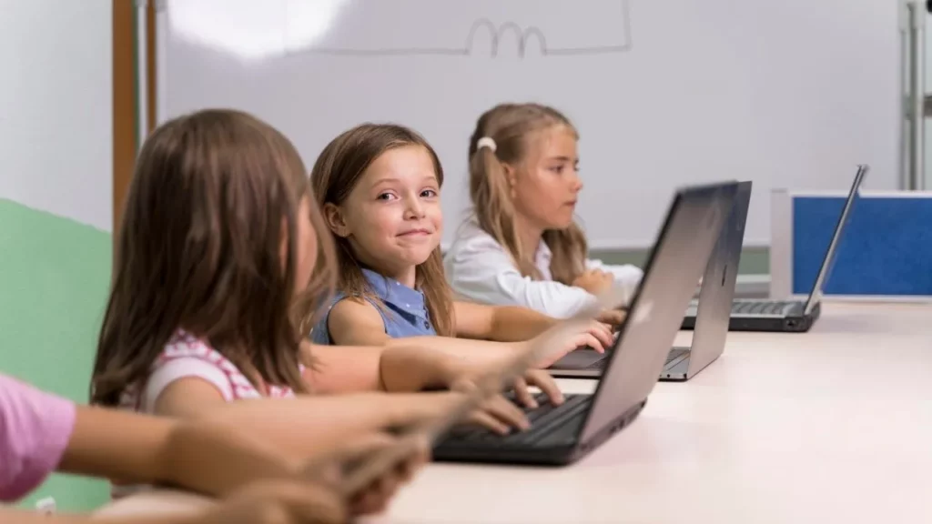 A group of smiling young girls (elementary school age) sit side by side at a long table in a bright classroom, happily working together on laptops with a colorful wall and whiteboard in the background.