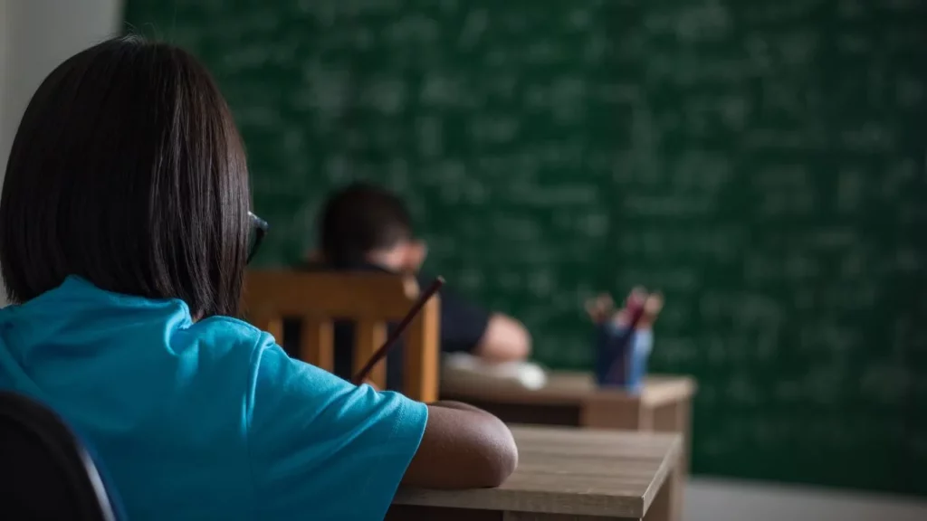 Girl from behind sitting at desk in classroom, facing a green chalkboard filled with writing.